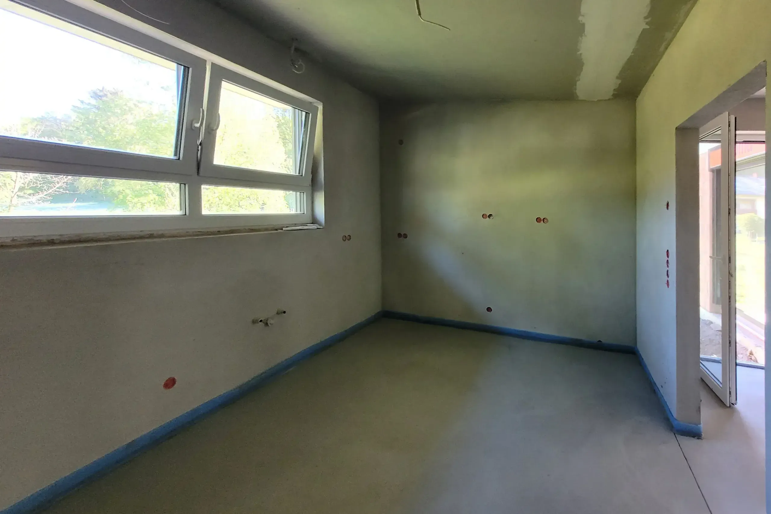 Rohbau-Aufnahme des verputzten Raums mit Fensterfront, Türöffnung und markierten Anschlusspunkten.