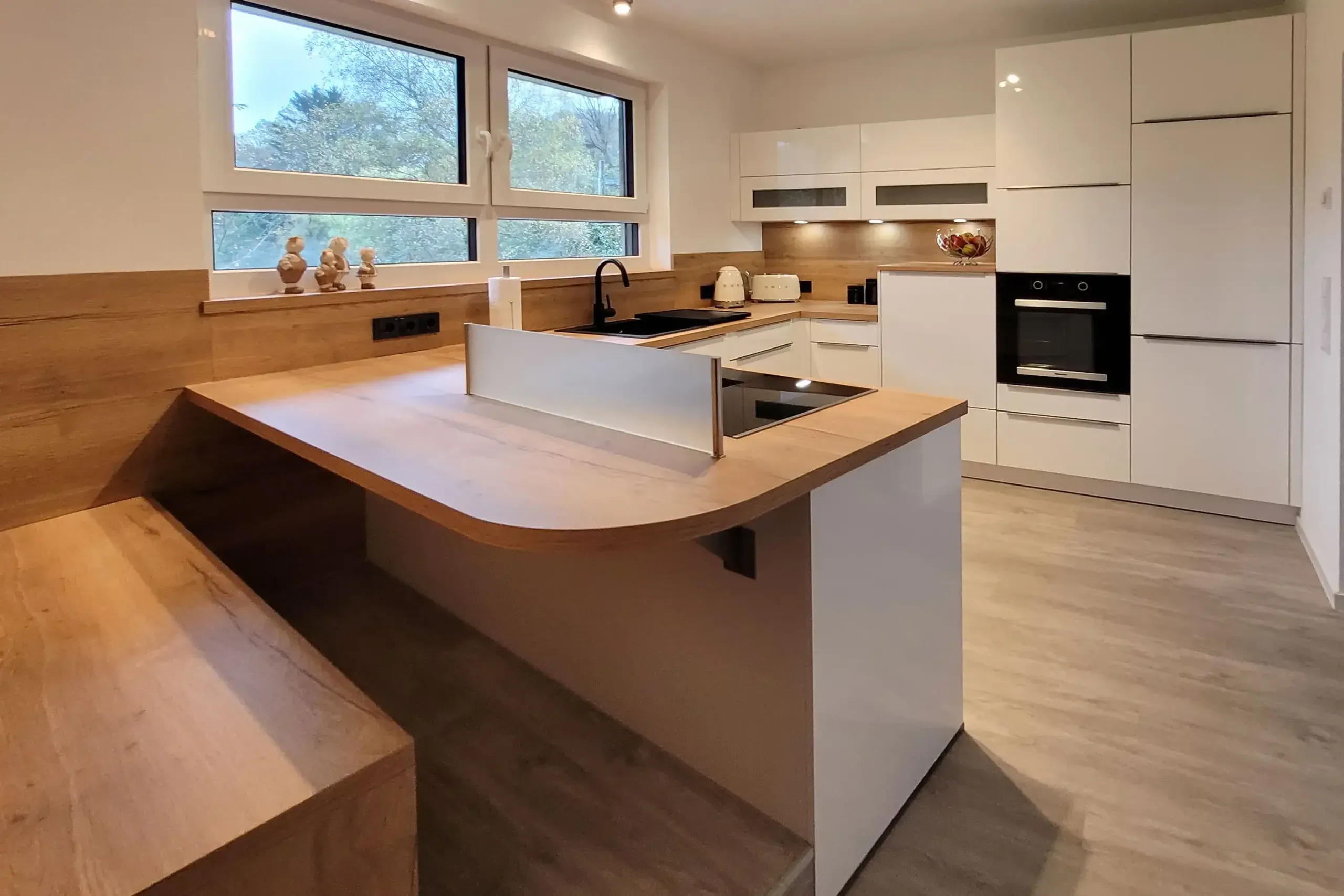 Weitwinkelansicht der weißen Hochglanzküche mit Halbinsel, Holz-Sitzbank und Wandzeile mit Backofen.