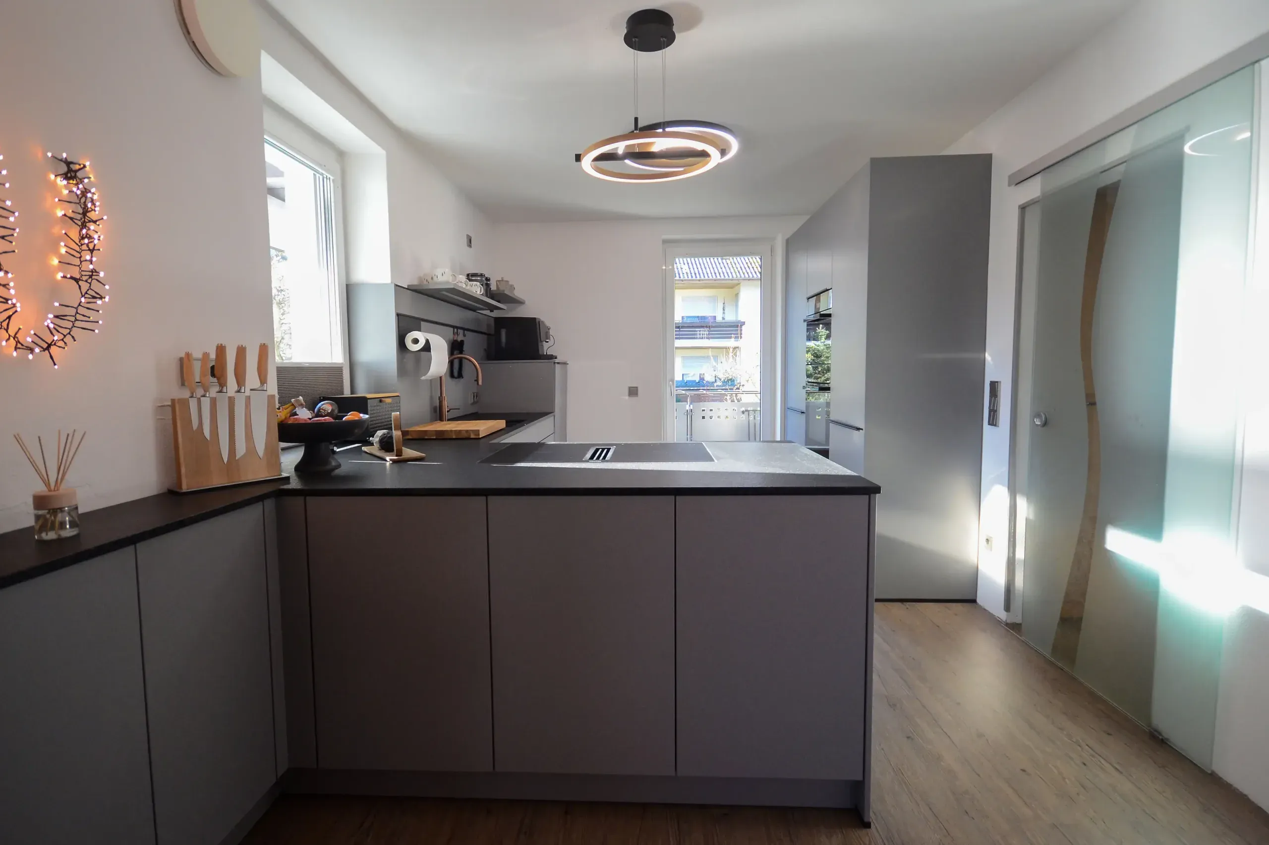 Längsperspektive aus dem Eingangsbereich auf die Halbinsel mit Muldenlüfter und schwarzer Naturstein-Arbeitsplatte, runde Doppel-LED-Pendelleuchte über der Insel, Fenster mit Messerblock links und Glasschiebetür mit Wellenmuster rechts – Ausstellungsküche im Broßler Küchenstudio Ehrbach