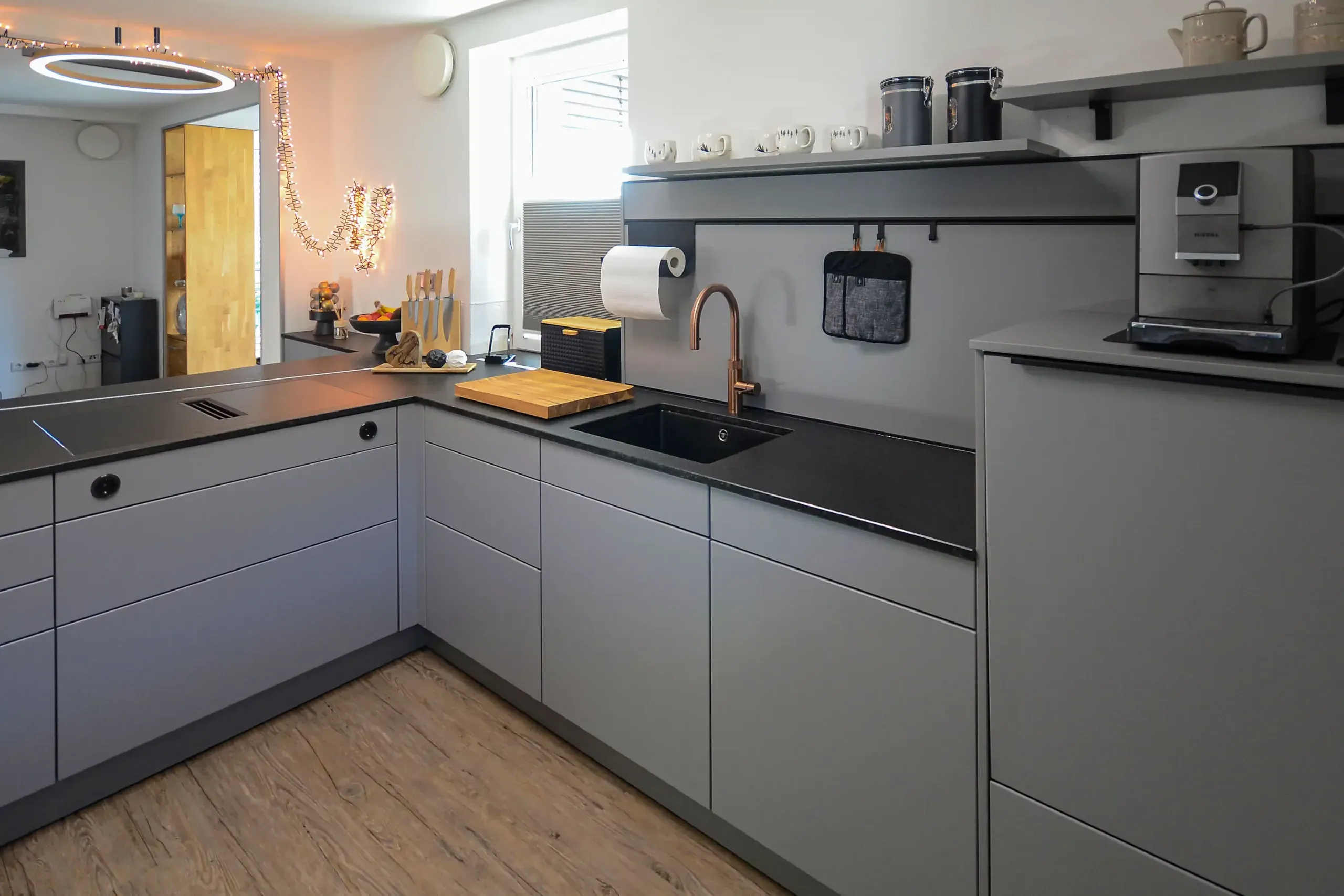 Moderne L-Küche mit grauen matten Fronten, schwarzer Naturstein-Arbeitsplatte, Spüle mit Kupferarmatur, runde LED-Pendelleuchte, Kaffeevollautomat, Wandregal mit Vorratsgläsern und Holzboden – Ausstellungsküche im Broßler Küchenstudio Ehrbach