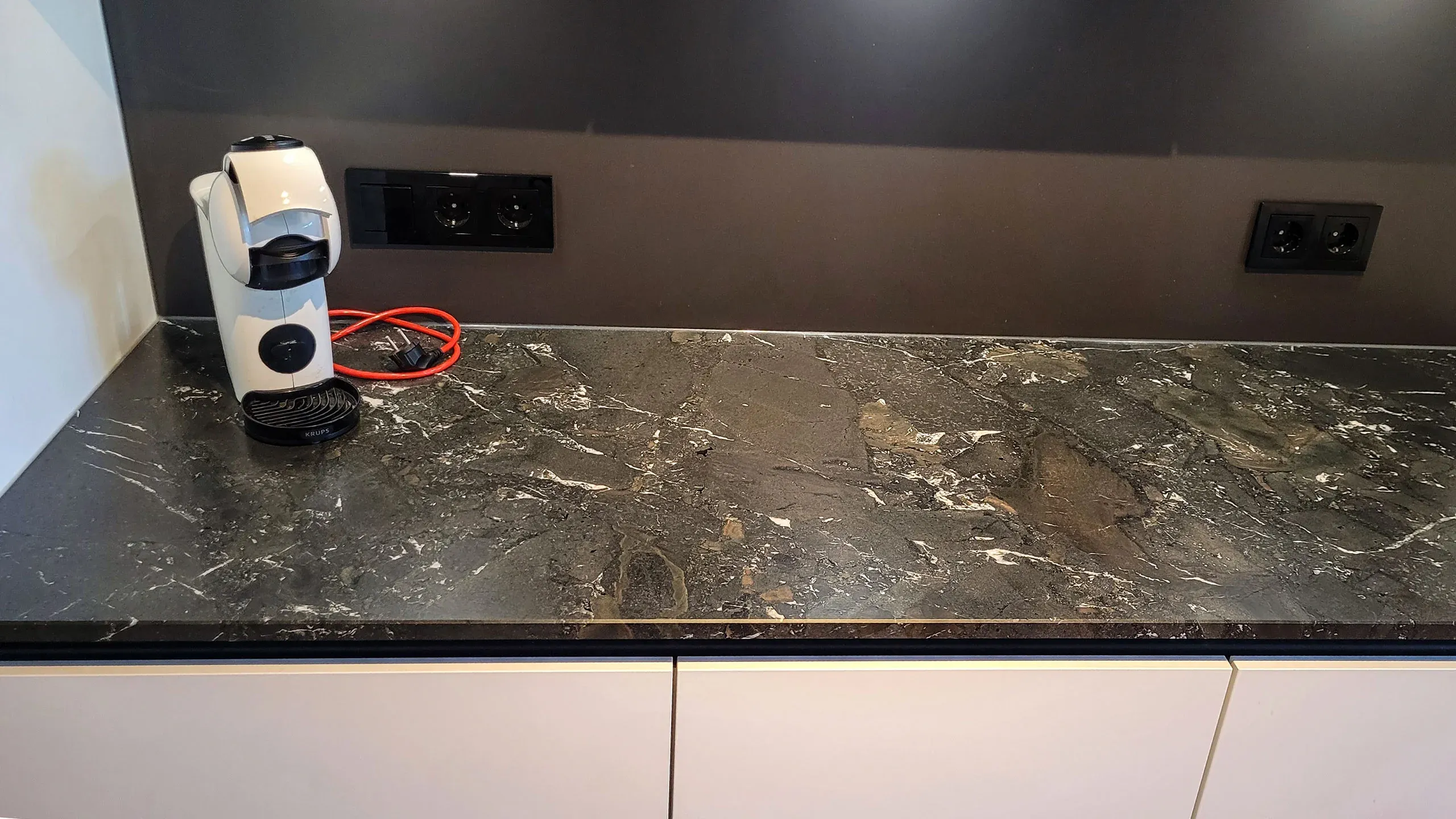 Detailansicht der dunklen Naturstein-Arbeitsplatte mit Kaffeekapselmaschine vor weißen Unterschränken.