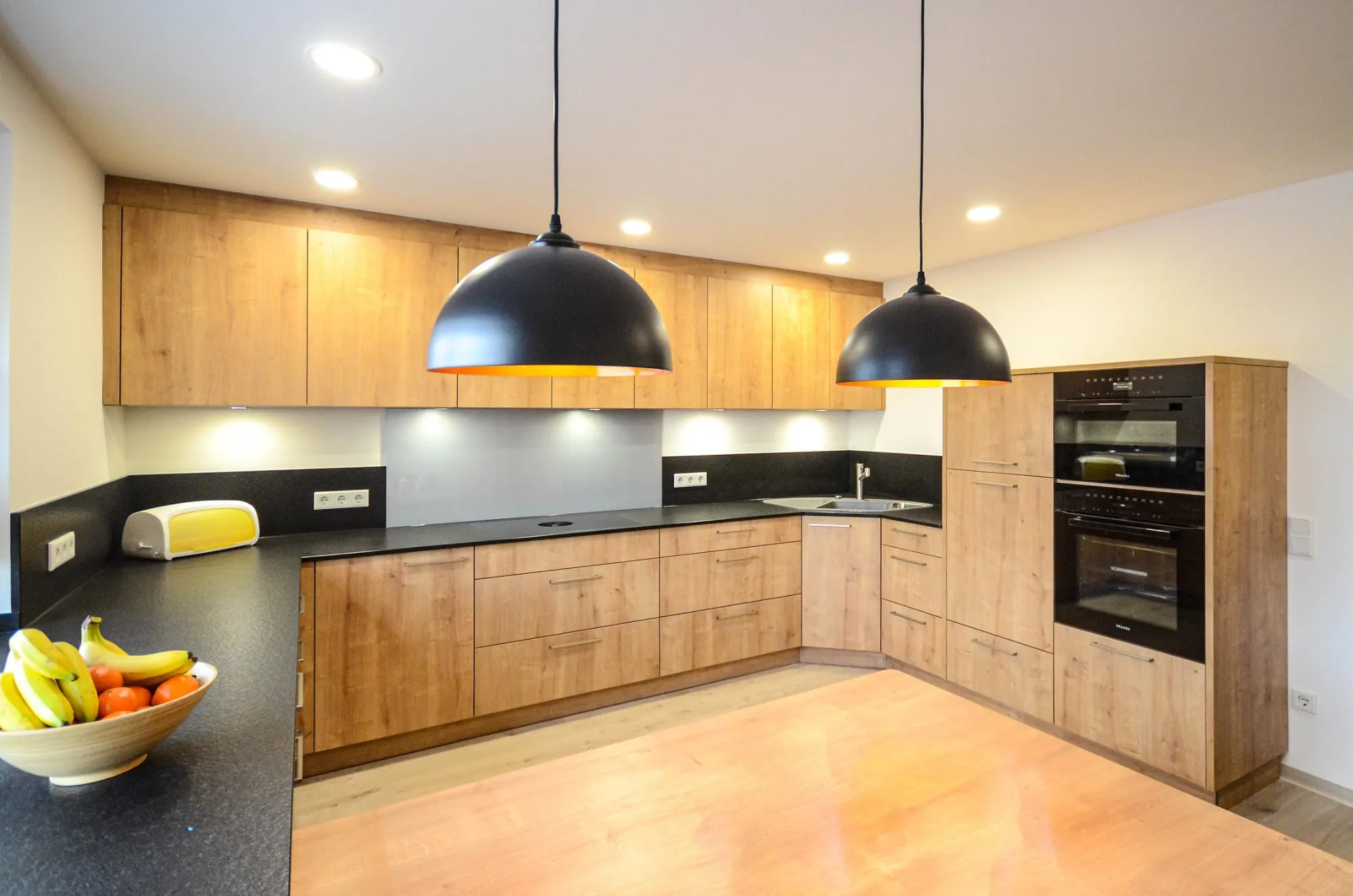 Nachher-Aufnahme: moderne L-Küche in Eichen-Holzoptik mit schwarzer Steinplatte, Hochschrank-Backofen und Kuppel-Pendelleuchten.