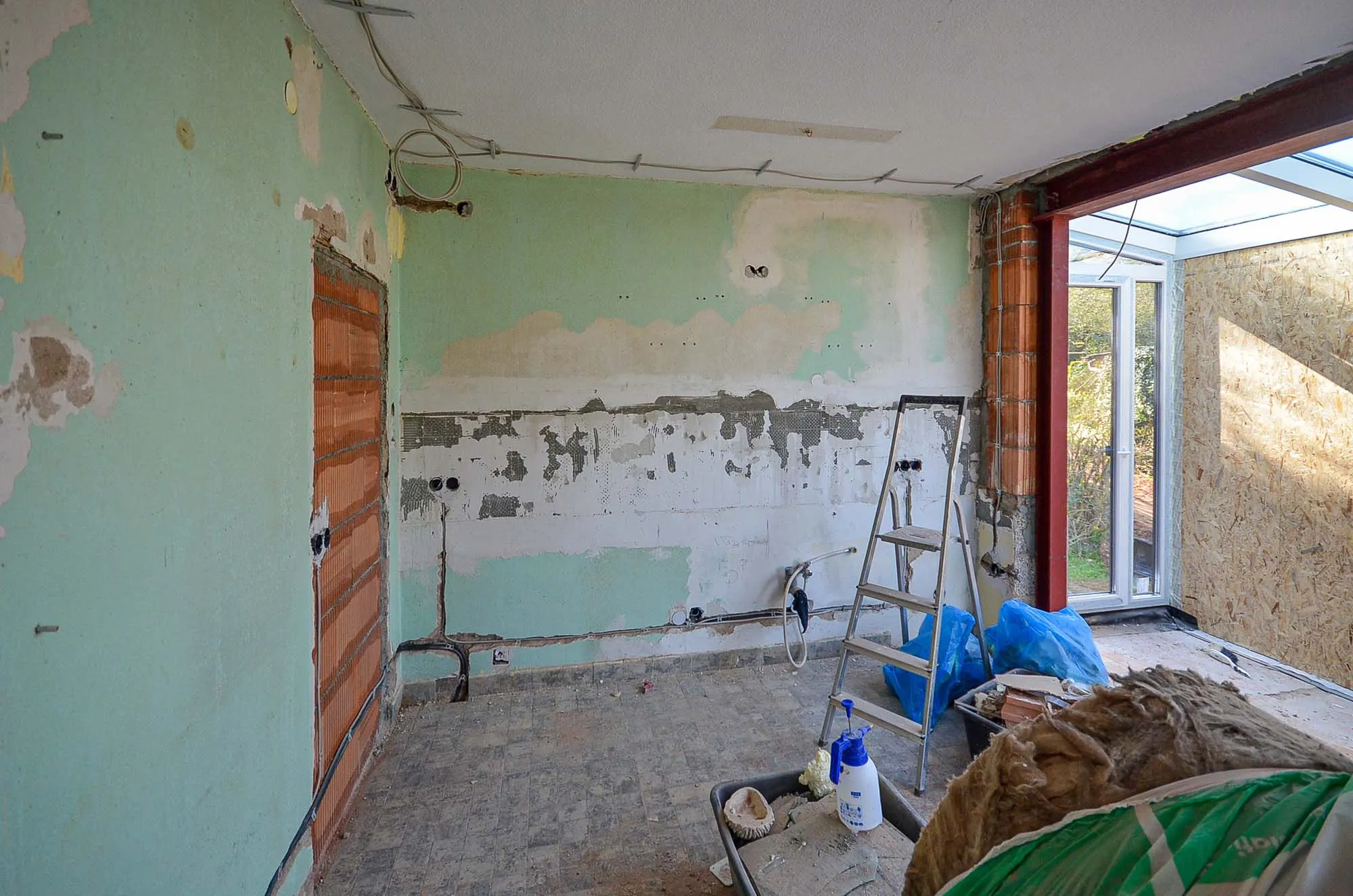 Vorher-Aufnahme der gegenüberliegenden Wand während der Entkernung, grüne Altwände mit teilweise abgeschlagenem Putz, sichtbare Elektroleitungen und freigelegtes Mauerwerk, Baumaterial im Vordergrund, rechts der neu eingebaute Wintergarten-Anbau mit Glasöffnung – Ausgangssituation für eine Küchenplanung von Dietmar Kraus bei Broßler Küche Aktiv