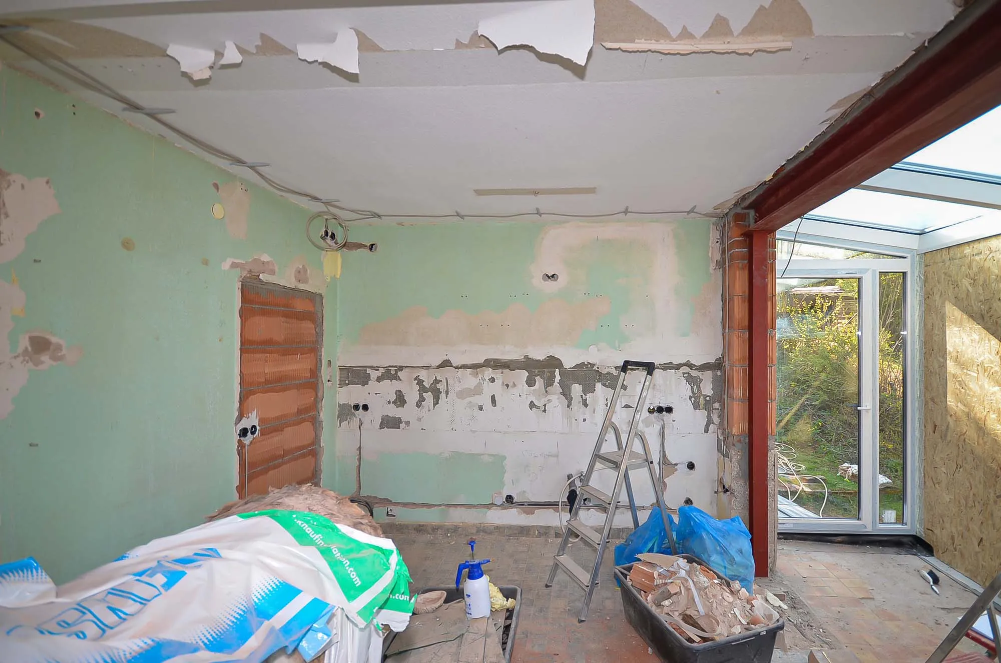 Vorher-Aufnahme während der Entkernung vor dem Kücheneinbau, grüne Altputz-Wände mit teilweise abgeschlagenen Fliesen, Baumaterial und Werkzeug im Raum, rechts ein neu eingebauter Wintergarten-Anbau mit großen Fenstern und Blick in den Garten – Ausgangssituation für eine Küchenplanung von Dietmar Kraus bei Broßler Küche Aktiv