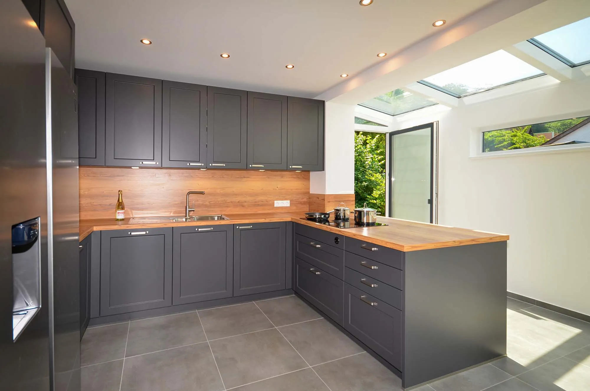 Moderne U-Küche in mattem Anthrazit mit Arbeitsplatte und Nischenrückwand in Eichen-Holzoptik, Edelstahl-Kühlschrank, lichtdurchfluteter Wintergarten-artiger Anbau mit Glasdach und Blick ins Grüne, grauer Fliesenboden – Küchenplanung von Dietmar Kraus für Broßler Küche Aktiv