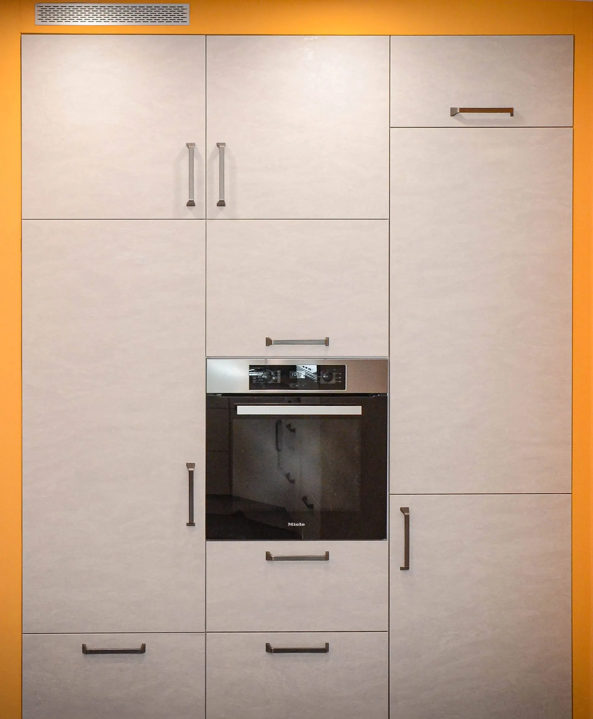 Detailaufnahme einer Hochschrankwand in beiger Steinoptik mit Bügelgriffen und integriertem Einbau-Backofen vor orangefarbener Wand.