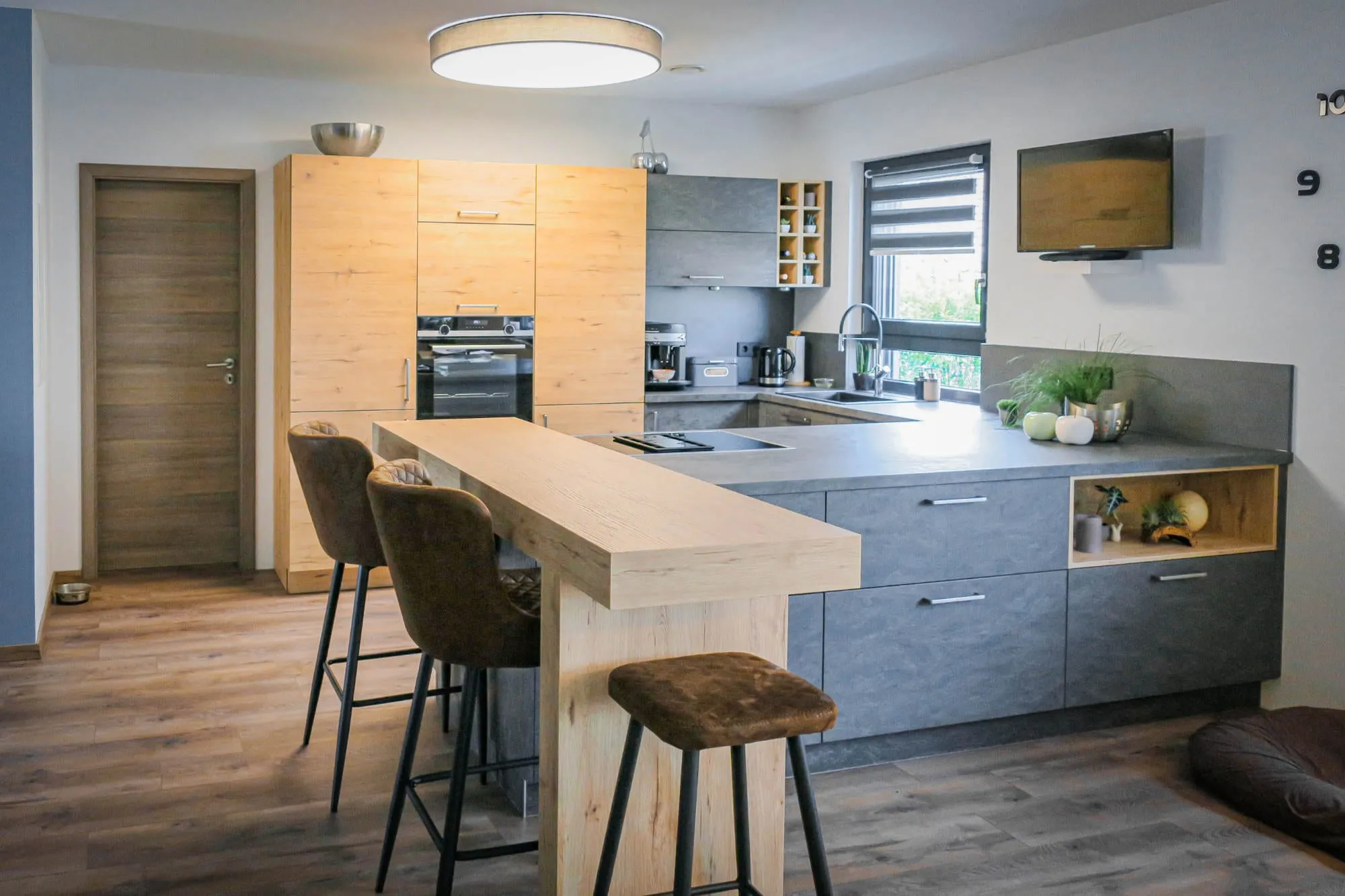 Halbinsel mit Holz-Bartheke und drei Cognac-Barhockern, im Hintergrund Küche mit anthrazitfarbenen Unterschränken, Eiche-Hochschränken und integriertem Holz-Nischenregal Holz grau