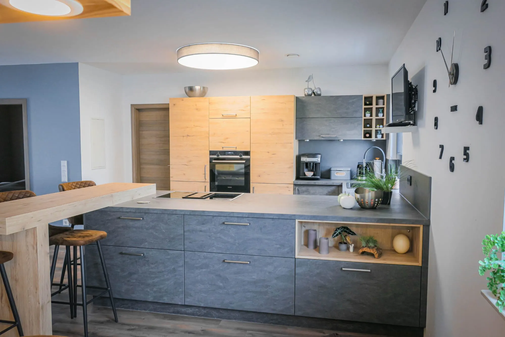 Offene Küche mit anthrazitfarbener Insel, integriertem Holz-Nischenregal und Eiche-Hochschränken, mit großer Wanduhr-Deko und runden Pendelleuchten Holz grau