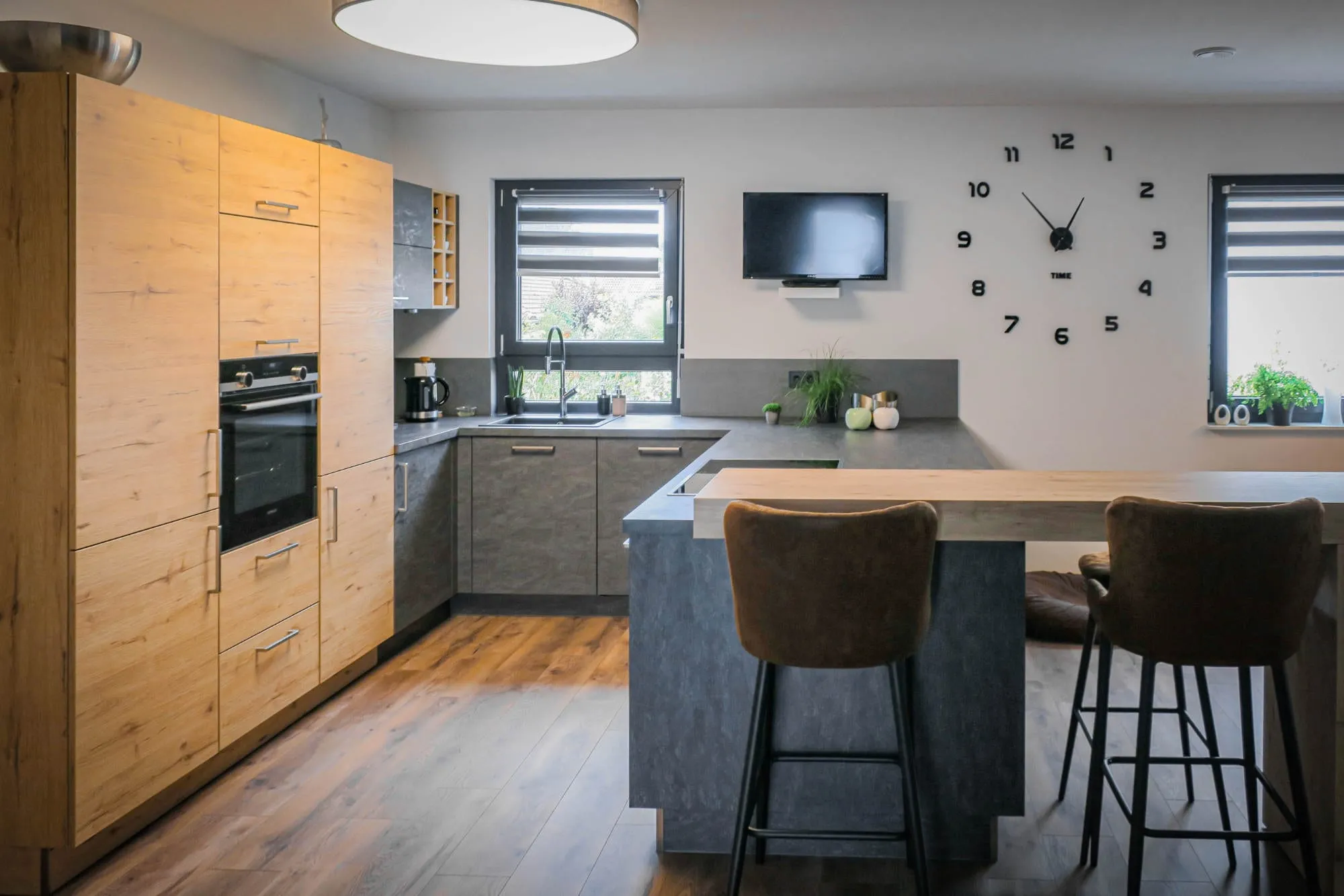U-Küche mit Holz-Hochschränken in Eiche-Optik und anthrazitfarbenen Unterschränken, mit erhöhter Bartheke aus Holz und zwei Cognac-Barhockern Holz grau