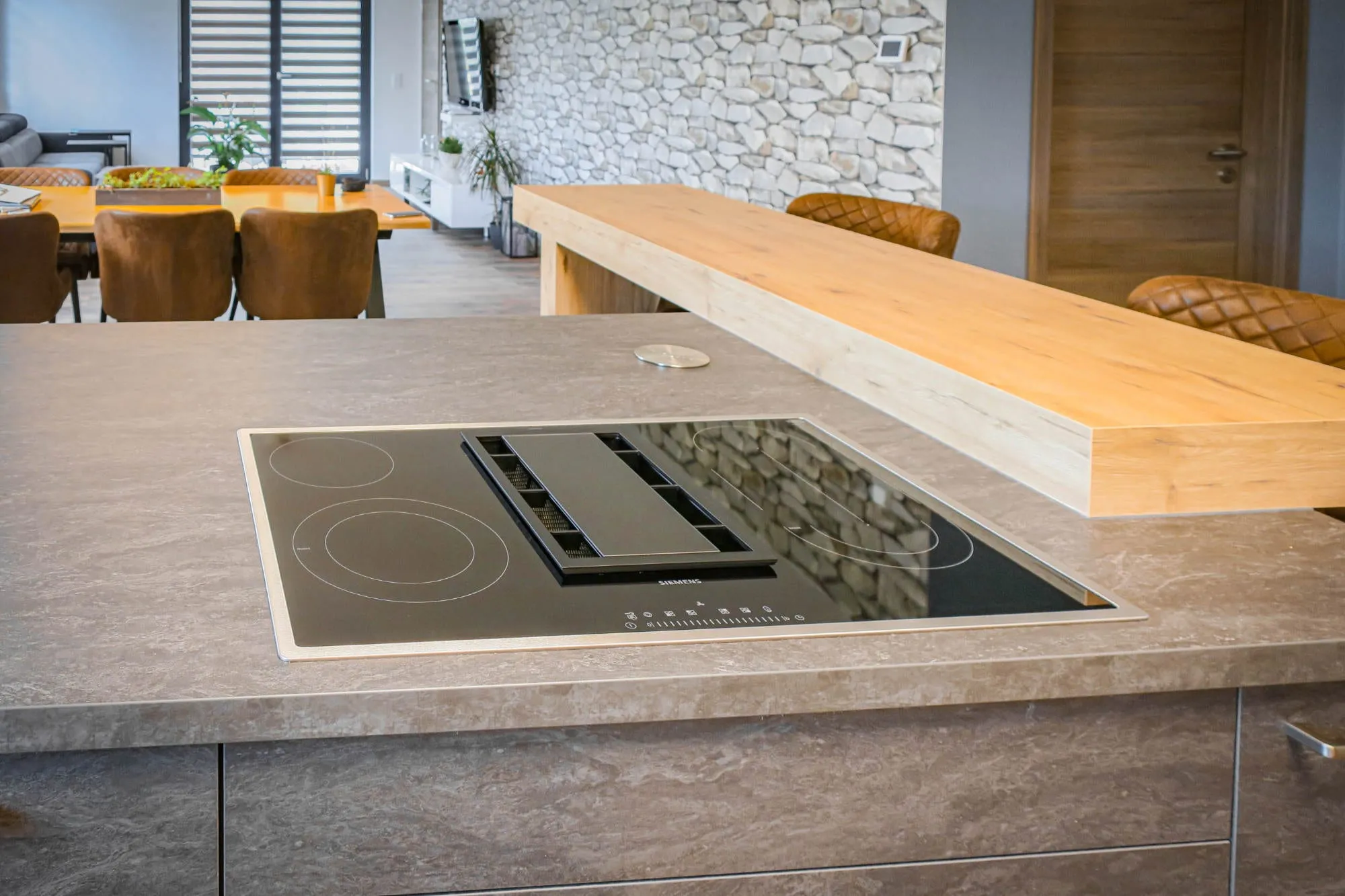 Detailaufnahme einer Kochinsel mit Steinoptik-Arbeitsplatte, Induktionskochfeld mit integriertem Muldenlüfter und erhöhter Holz-Bartheke vor einer Bruchsteinwand Holz grau