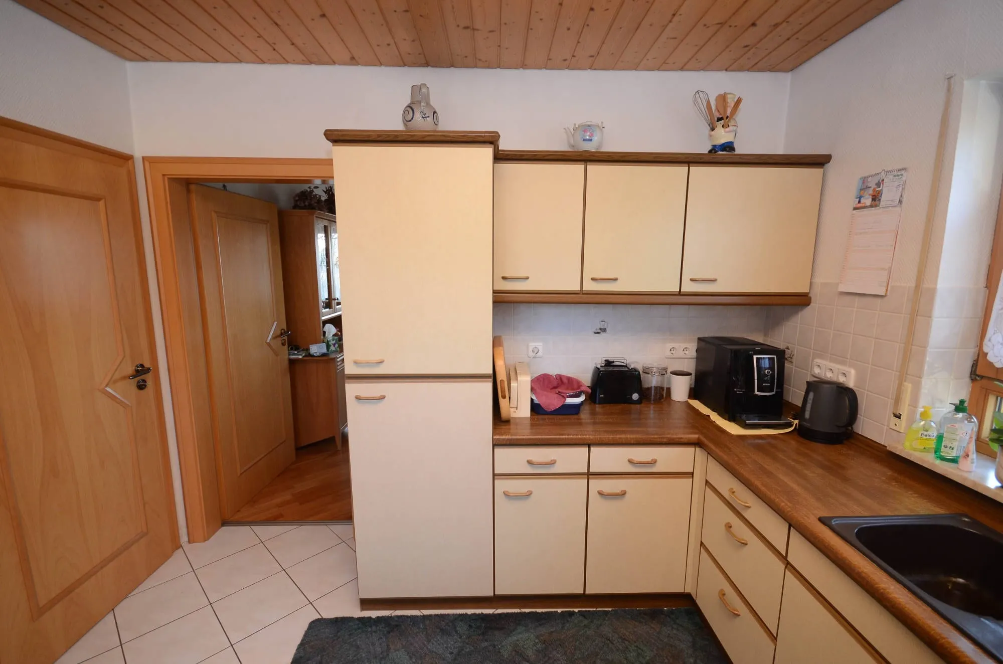 Vorher-Aufnahme einer älteren L-Küche in beiger Holzoptik mit Holzarbeitsplatte und Holzdecke.