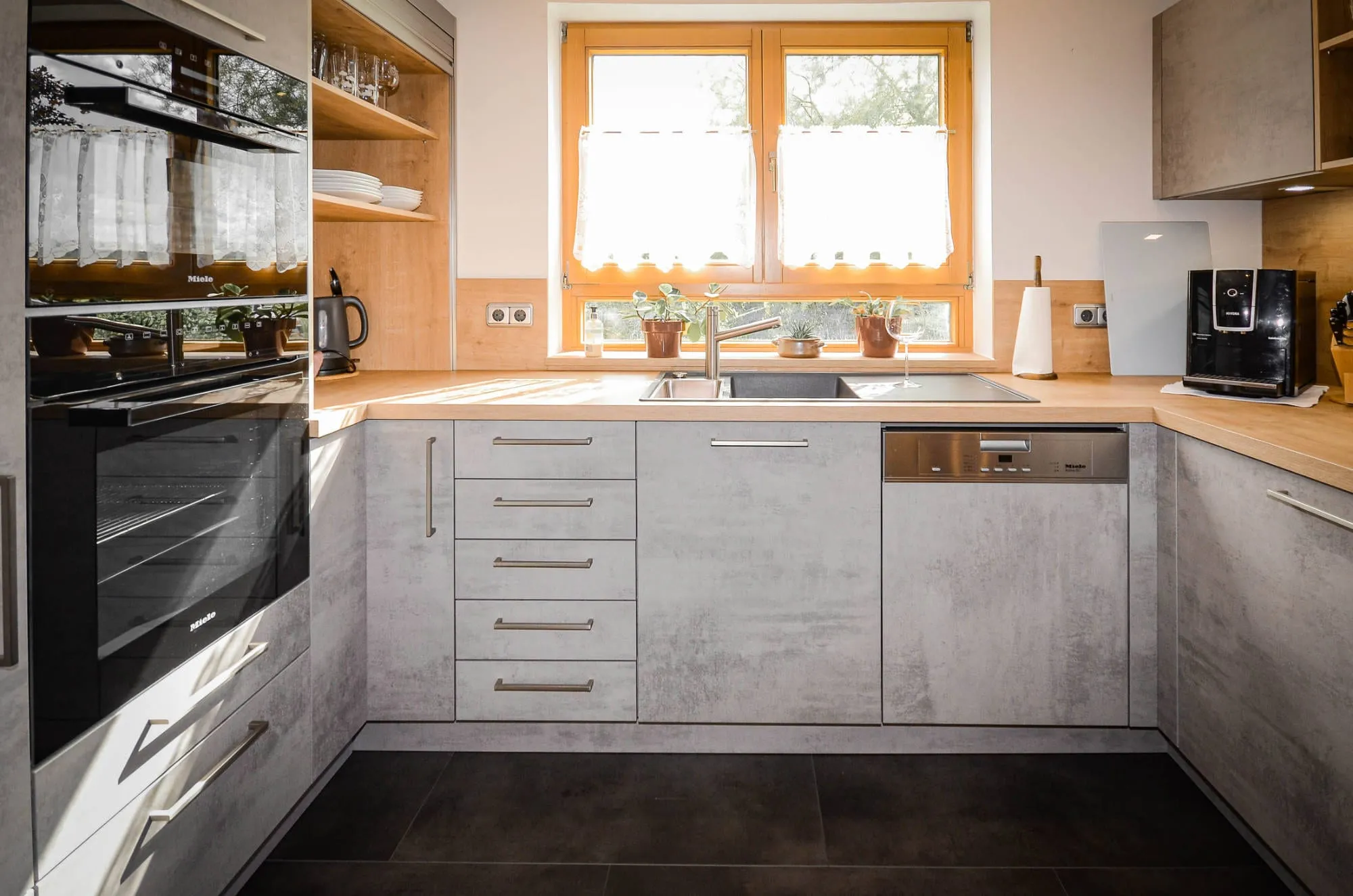 Frontalansicht der U-Küche in Beton-Steinoptik mit Spüle unter Doppelfenster und seitlichem Hochschrank mit Einbau-Backofen.