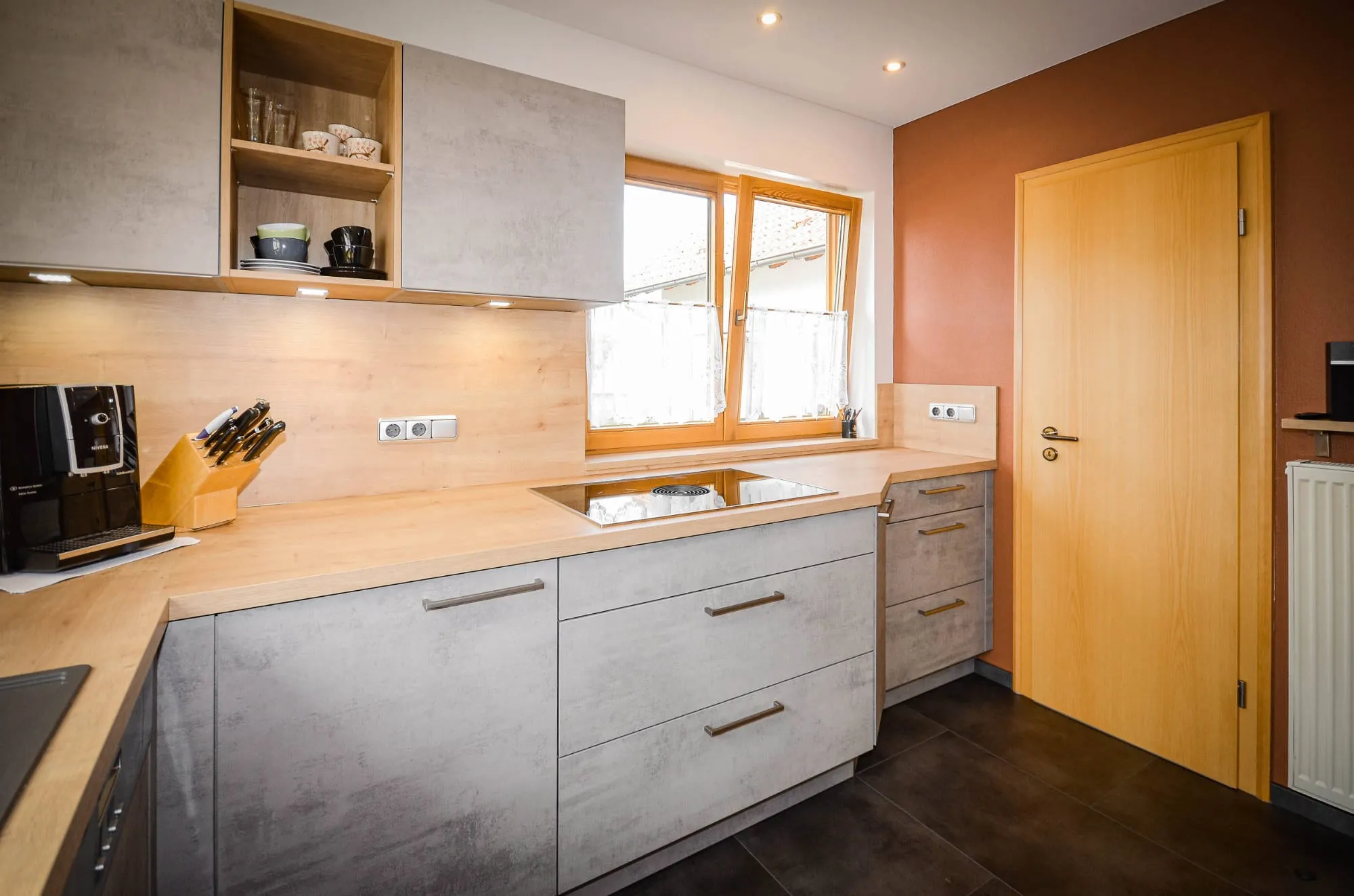 U-Küche in heller Beton-Steinoptik mit Massivholz-Arbeitsplatte, Induktionskochfeld unter dem Fenster und offenem Holz-Wandregal.