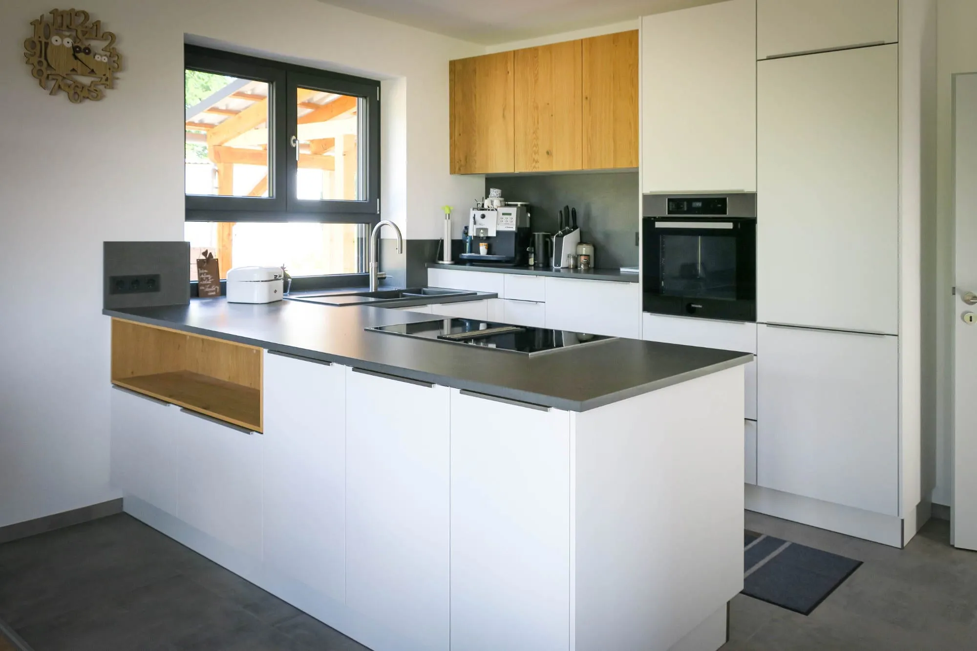 Offene moderne Küche mit weißen grifflosen Fronten, anthrazitfarbener Arbeitsplatte, Halbinsel mit Induktionskochfeld und seitlichem offenen Holz-Nischenregal