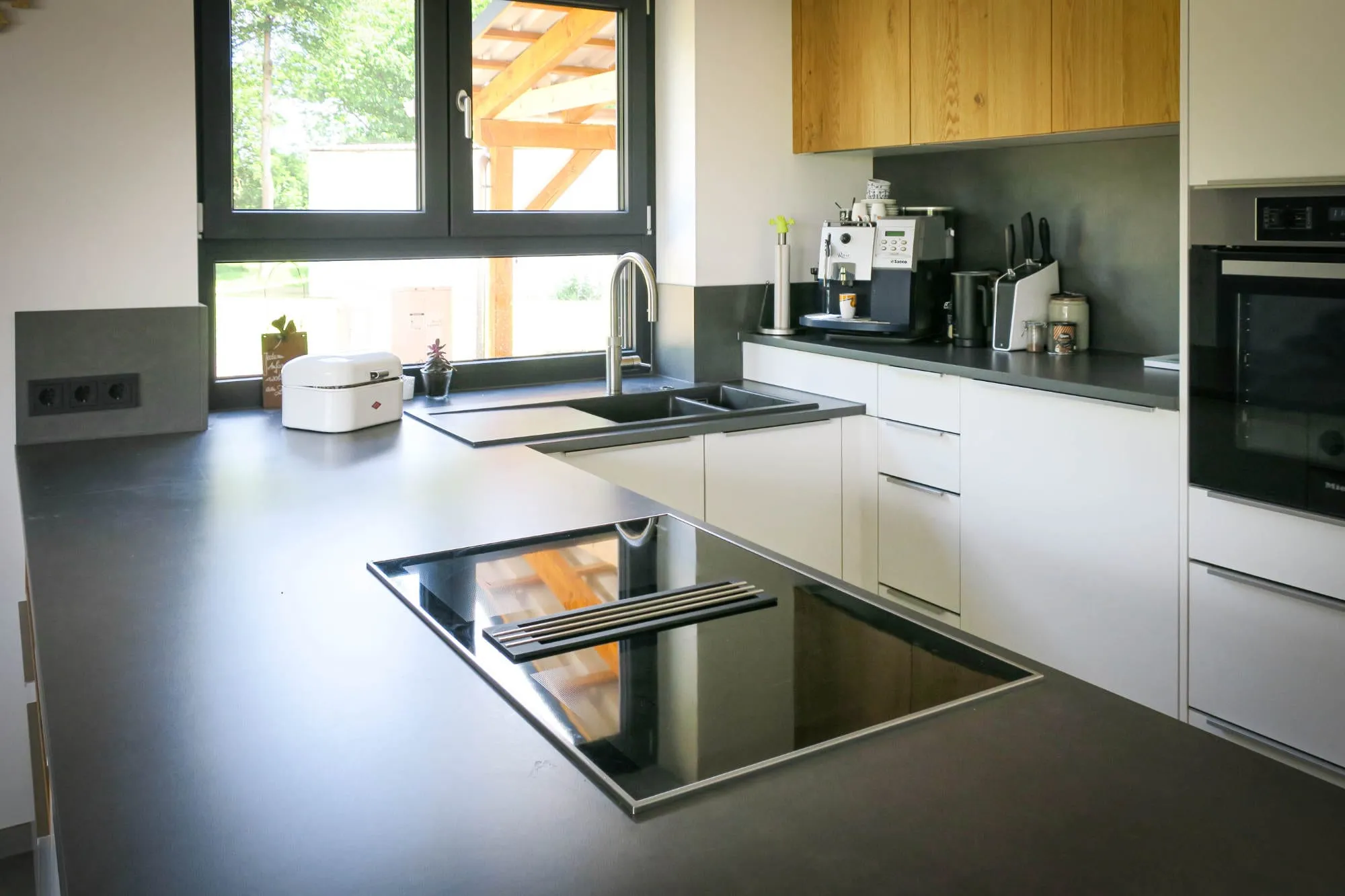 Edelstahl-Kochinsel mit eingelassenem Teppanyaki-Grill, im Hintergrund Doppelspüle am Fenster, weiße Unterschränke und Eiche-Hochschränke modern