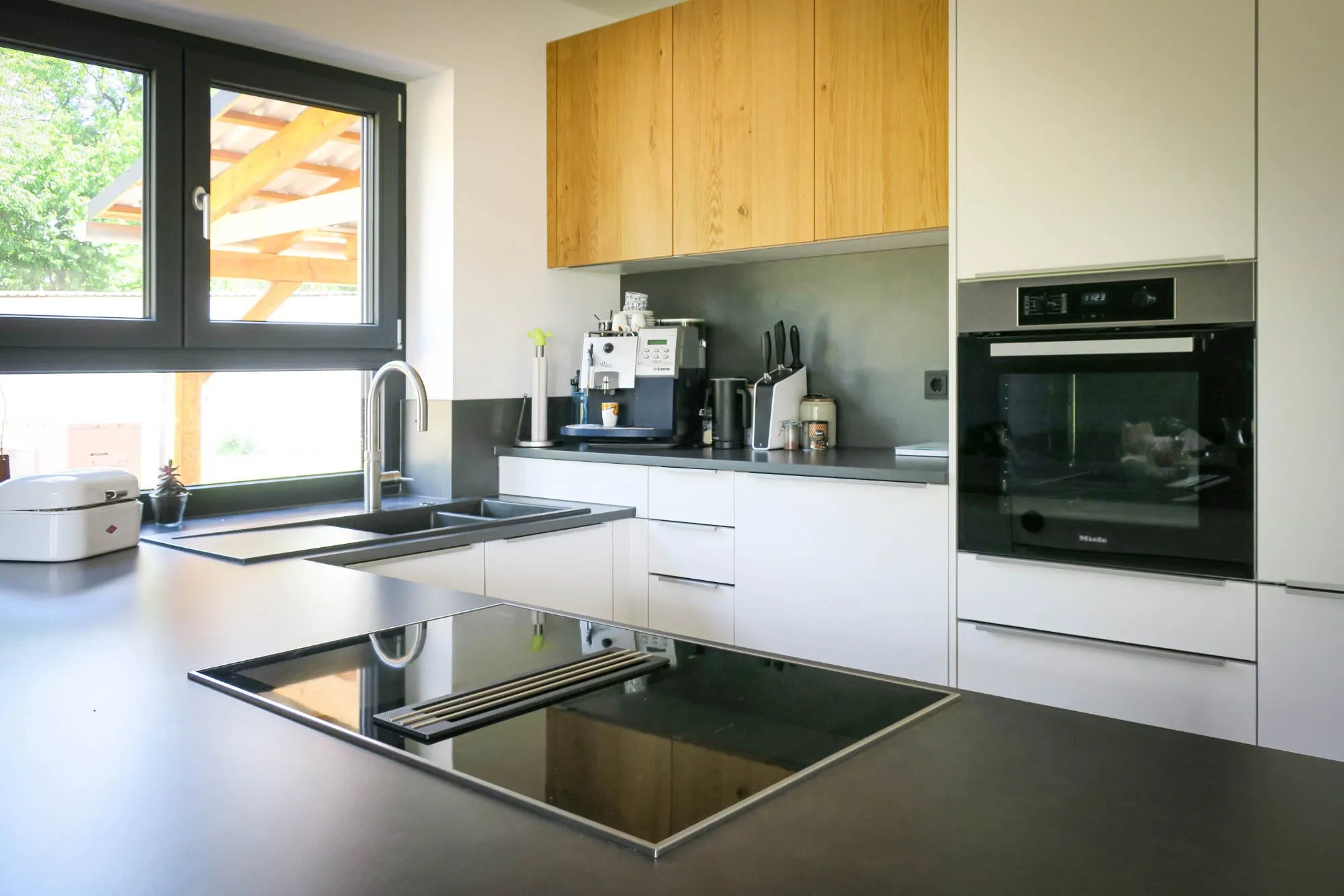Moderne grifflose Küche mit weißen Hochglanzfronten, Eiche-Hochschränken und Edelstahl-Arbeitsplatte, mit Doppelspüle am Fenster und Edelstahl-Kochinsel mit integriertem Teppanyaki-Grill modern