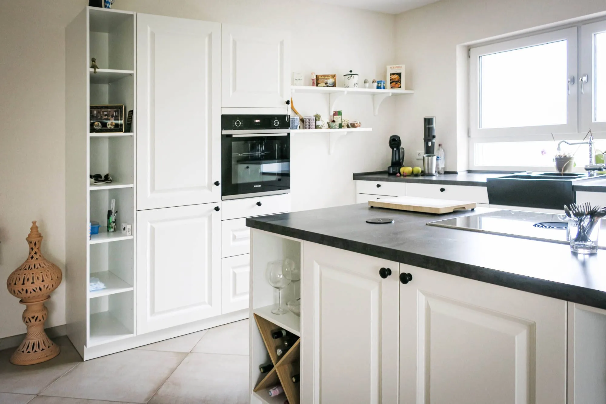 Weiße Landhausküche mit Backofen-Hochschrank, freistehendem offenen Regal, Kochinsel mit Weinregal und Spüle am Fenster