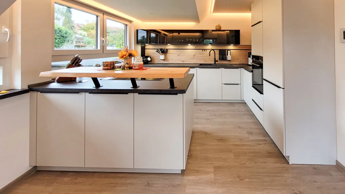 Offene weiße Küche mit Holz-Bartheke, dunkler Arbeitsplatte und Backofen-Hochschrank vor Fensterfront.