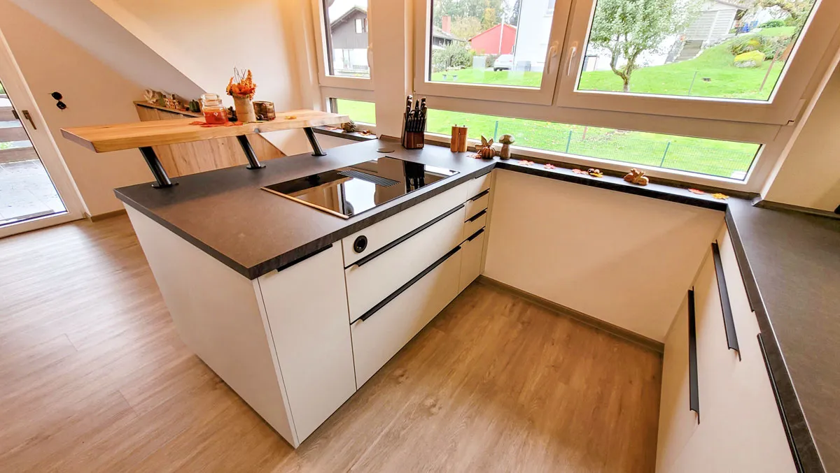 Weiße Kochinsel mit Muldenlüfter und angesetzter Holz-Bartheke vor einem Eckfenster mit Gartenblick.