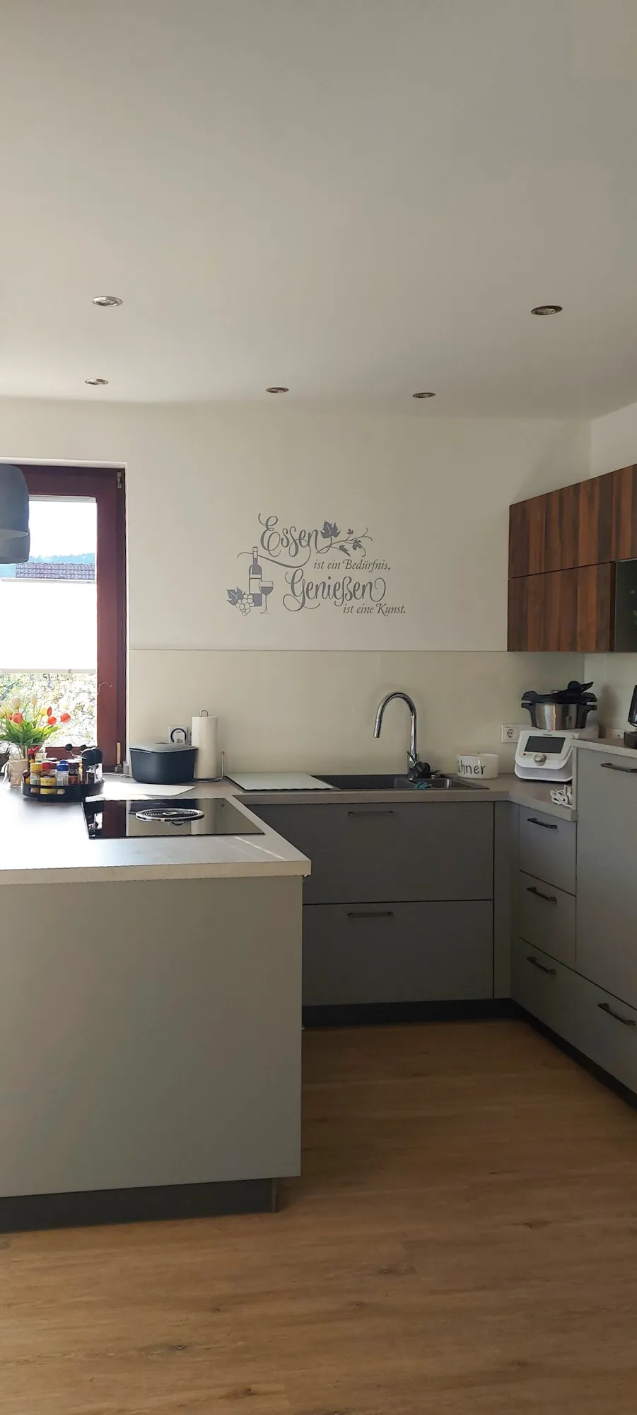 Graue L-Küche mit Halbinsel, Kochfeld, Holz-Hochschrank und Wandspruch-Deko